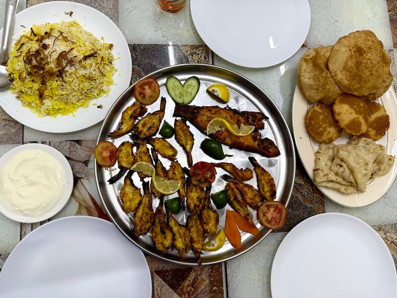 A colourful meal spread featuring grilled prawns and fish garnished with lemon slices, alongside biryani topped with fried onions, fluffy puris, flatbread, and a bowl of yoghurt. The dishes are neatly arranged on a tiled table, ready to be served.