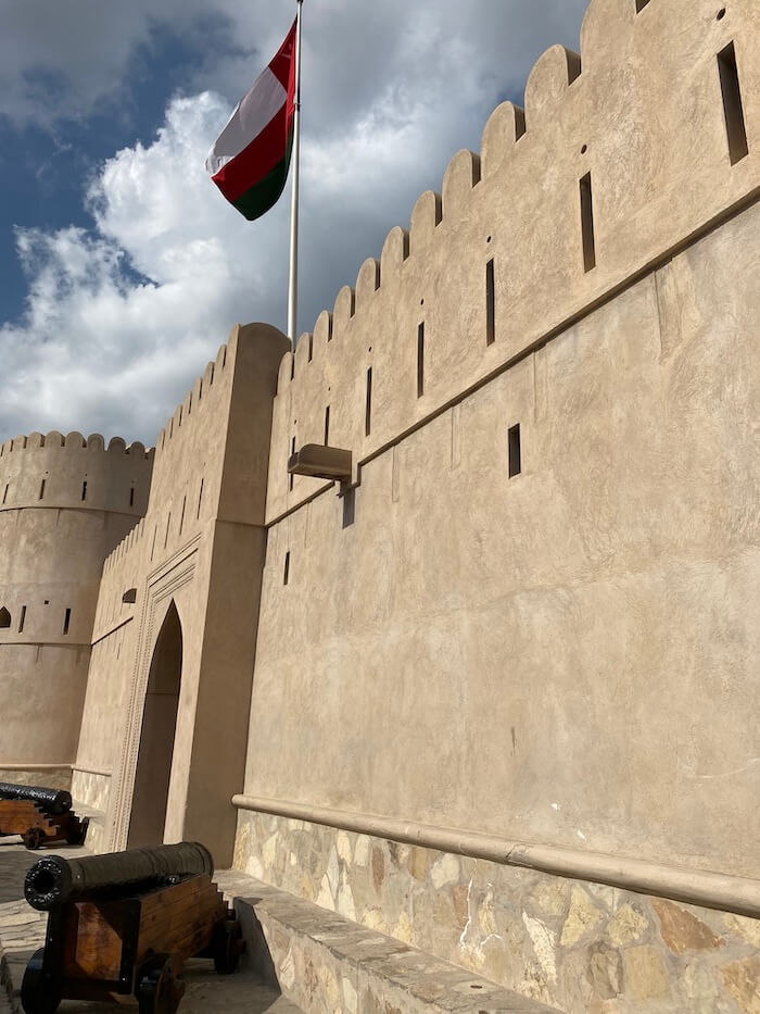 A beige stone fort wall with battlements stretches under a partly cloudy sky, with the Omani flag flying high above and cannons lined up along the pathway.