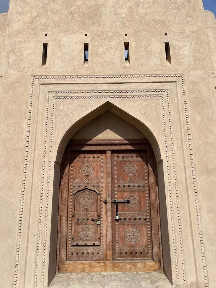 A large wooden door with intricate geometric carvings and metal studs is set within a pointed archway of a sand-coloured fort wall. A padlock secures the door, and a smaller inset door is visible within it.