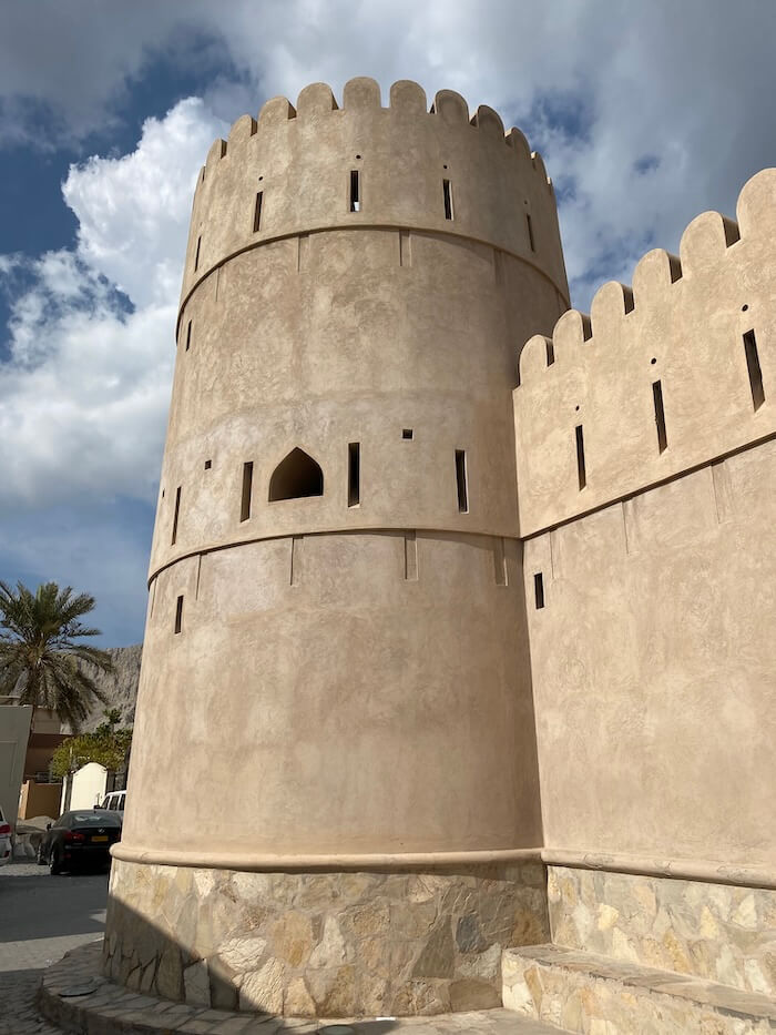 A rounded watchtower rises from the corner of the fort, with narrow vertical slits and arched windows, built from the same sandy-hued material as the walls.