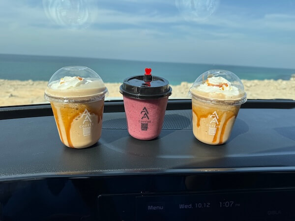Three iced drinks from AFA Coffee are placed on a car dashboard with a beach and sea view in the background. Two are caramel frappes topped with whipped cream, and the middle one is a pink smoothie with a heart-shaped stirrer.