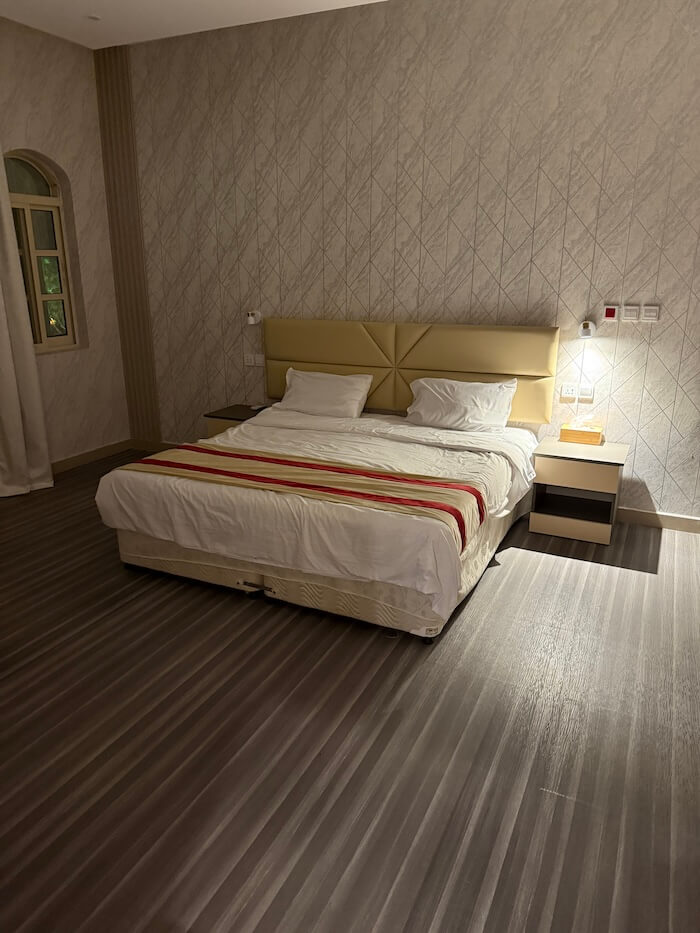 A double bed with a padded headboard sits in a spacious hotel room with patterned wallpaper and striped flooring. A soft bedside lamp casts a warm glow over the room.