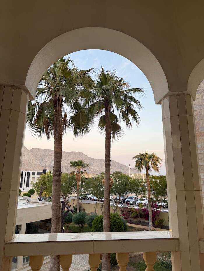 View from a balcony framed by arched pillars, showing tall palm trees, manicured gardens, parked cars, and rocky mountains in the distance under a pastel evening sky.