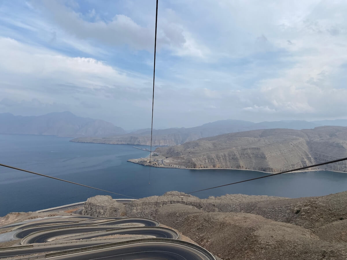 Winding mountain roads curve dramatically down towards a calm blue sea, with rugged cliffs and hazy mountains in the distance. Zipline cables stretch across the foreground, suggesting a scenic adventure viewpoint.