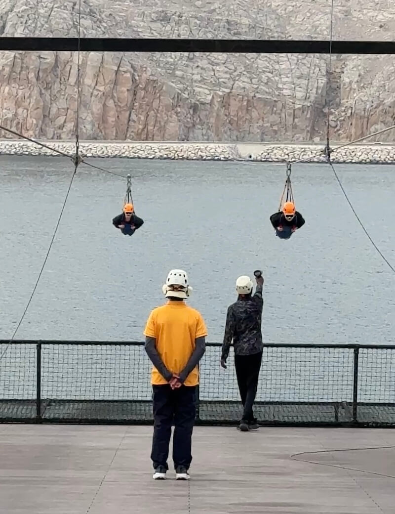 Two zipliners in flight position are suspended over water, moments before launch, while two staff members in helmets observe and signal from the platform. Behind them, rocky cliffs and a calm sea set a dramatic backdrop.