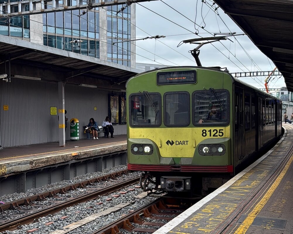 A green DART (Dublin Area Rapid Transit) train numbered 8125 arrives at a covered platform with overhead electric lines at a city station. Two passengers sit on a bench to the left, and the platform markings and surrounding urban architecture are clearly visible.