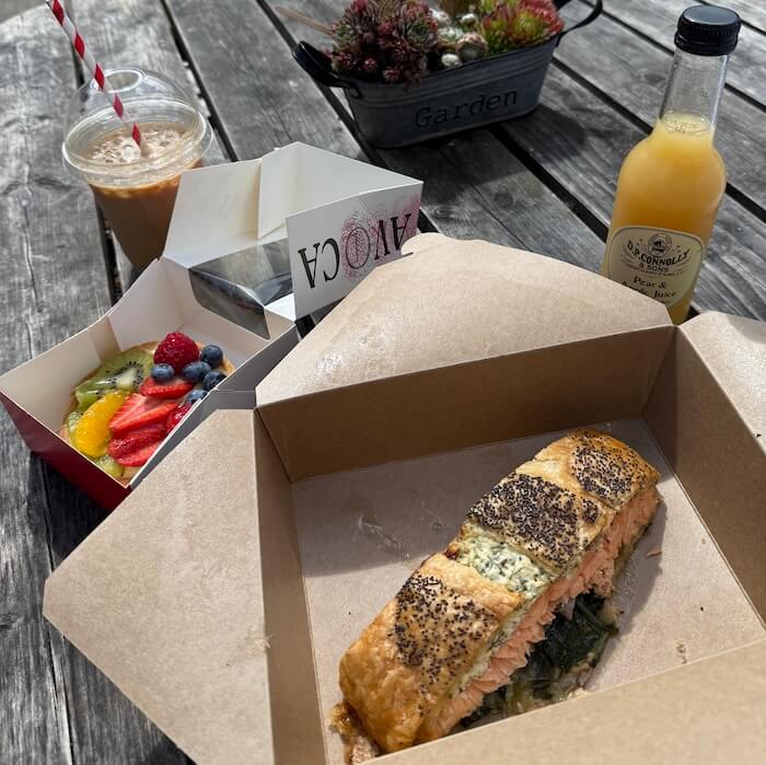 Outdoor lunch on a rustic wooden table featuring a box with a slice of salmon en croute topped with poppy seeds, a fruit tart with fresh kiwi, strawberries, blueberries, and orange slices, a bottle of pear and apple juice, and an iced coffee with a red striped straw. A small planter labelled "Garden" adds a decorative touch in the background.