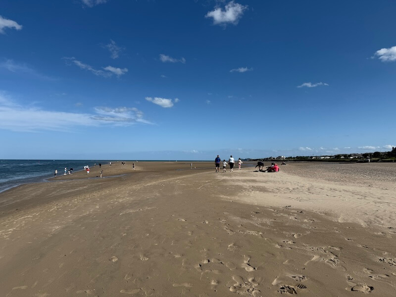 Wide sandy beach under a clear blue sky with scattered clouds, where people are walking and relaxing near the shoreline. The calm sea stretches along the left while a row of houses is visible in the distance to the right.