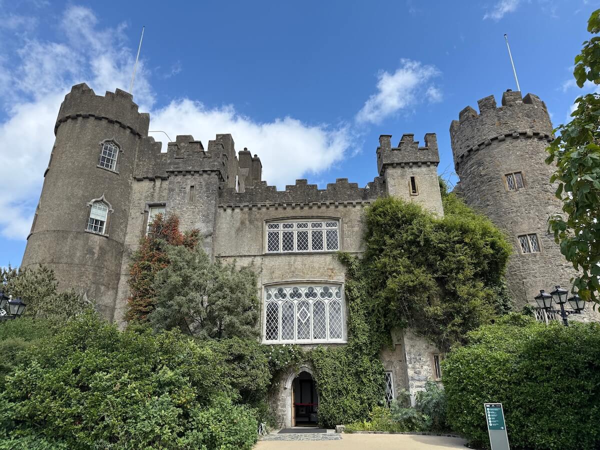 Front view of Malahide Castle in Dublin, Ireland, featuring grey stone walls, crenellated towers, and arched lattice windows surrounded by lush green ivy and trees. The blue sky and soft clouds frame the historic architecture, enhancing its medieval charm.