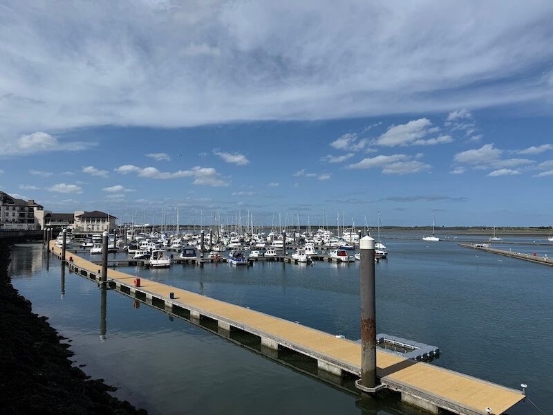 Marina filled with white boats and yachts docked at floating yellow piers, set against a calm blue harbor under a partly cloudy sky. Waterfront buildings sit on the left, framing the peaceful coastal scene.