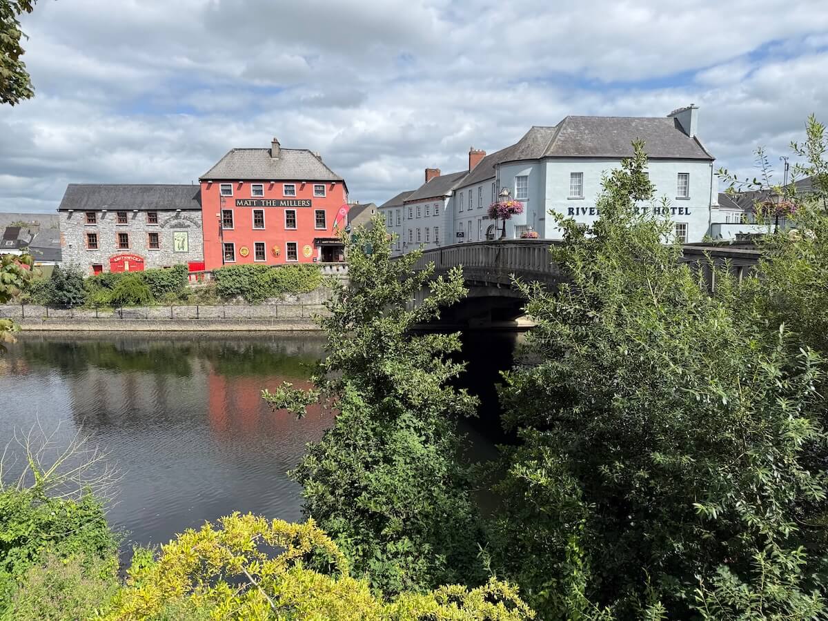 A riverside view shows colourful buildings including the bright red “Matt the Millers” pub and the pale blue River Court Hotel, with their reflections shimmering on the water beneath a cloudy sky.