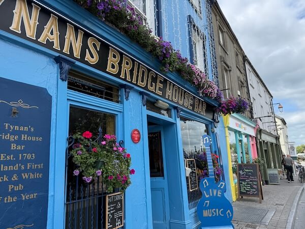A bright blue pub with gold signage, flower baskets, and a “Live Music” sign on the pavement, with other colourful shopfronts along the street.