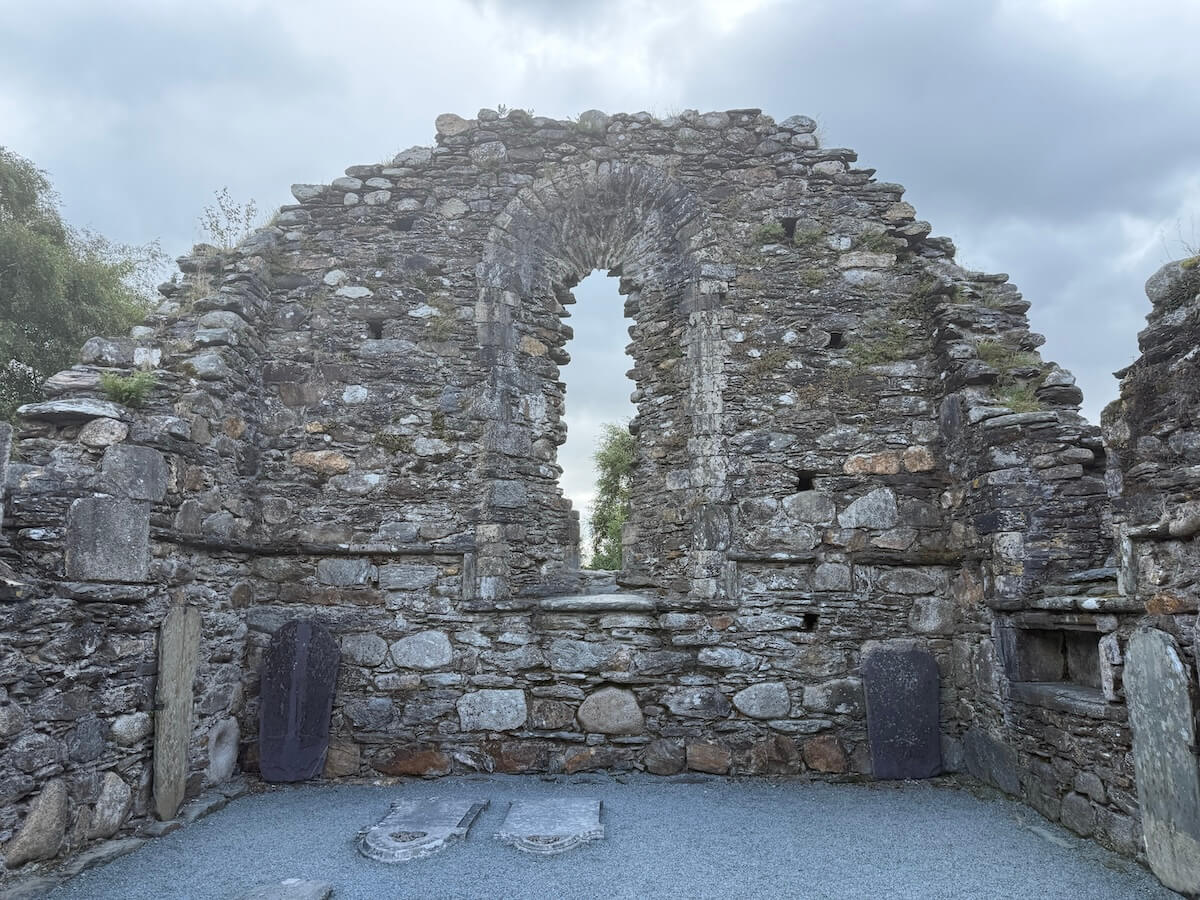 The weathered stone ruins of a medieval church wall feature a tall arched window opening with no glass, framed by rough masonry. Gravestones and stone slabs rest at the base, with cloudy skies adding to the solemn atmosphere.
