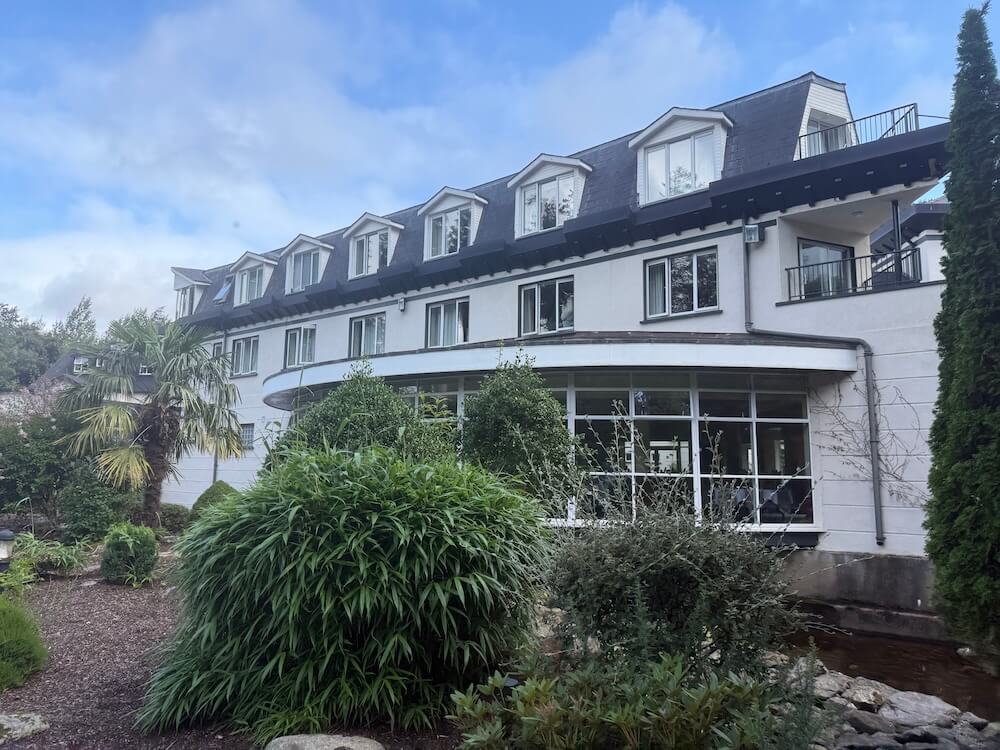 A modern white hotel with dormer windows and a sloping dark roof stands behind landscaped gardens filled with palms, tall shrubs, and ornamental greenery under a partly cloudy blue sky.
