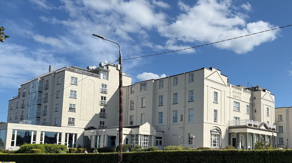 The Grand Hotel, a large cream-colored historic building with multiple stories and traditional architectural details, stands under a blue sky with scattered clouds and is fronted by trimmed hedges and a glass entryway.
