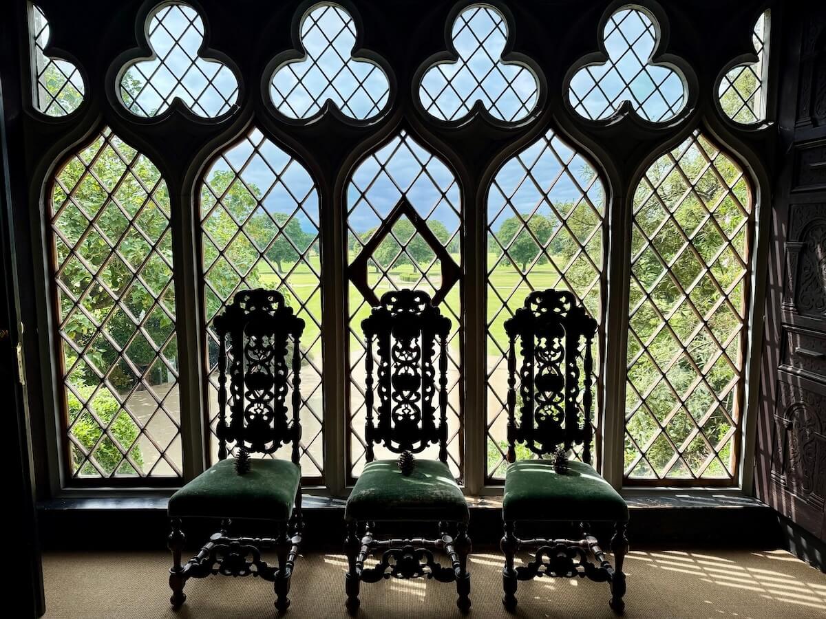 Three ornate wooden chairs with green velvet seats are placed in front of tall, arched windows with diamond-patterned glass panes. The windows frame a view of lush green gardens and distant fields under a cloudy sky.