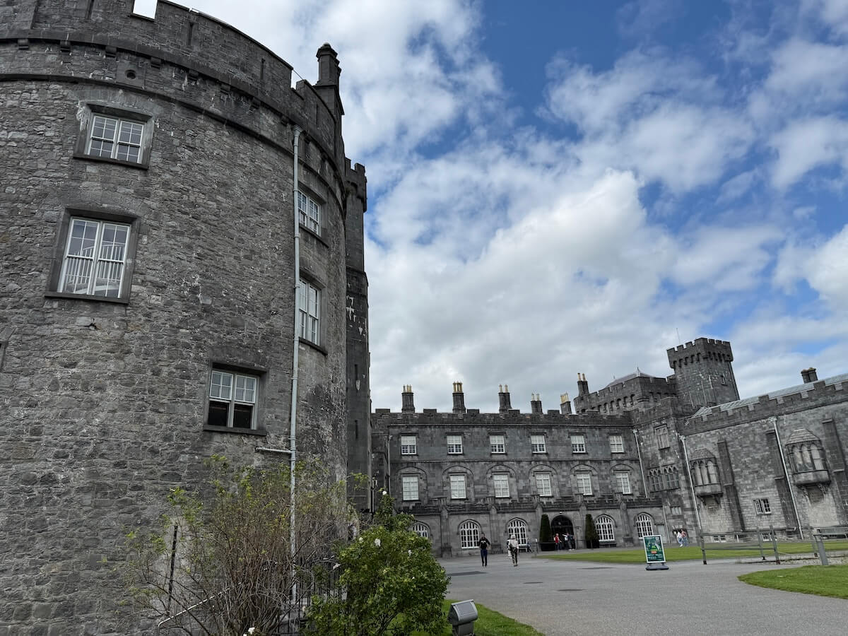 The stone walls and towers of Kilkenny Castle rise against a partly cloudy sky, with visitors walking in the courtyard below.