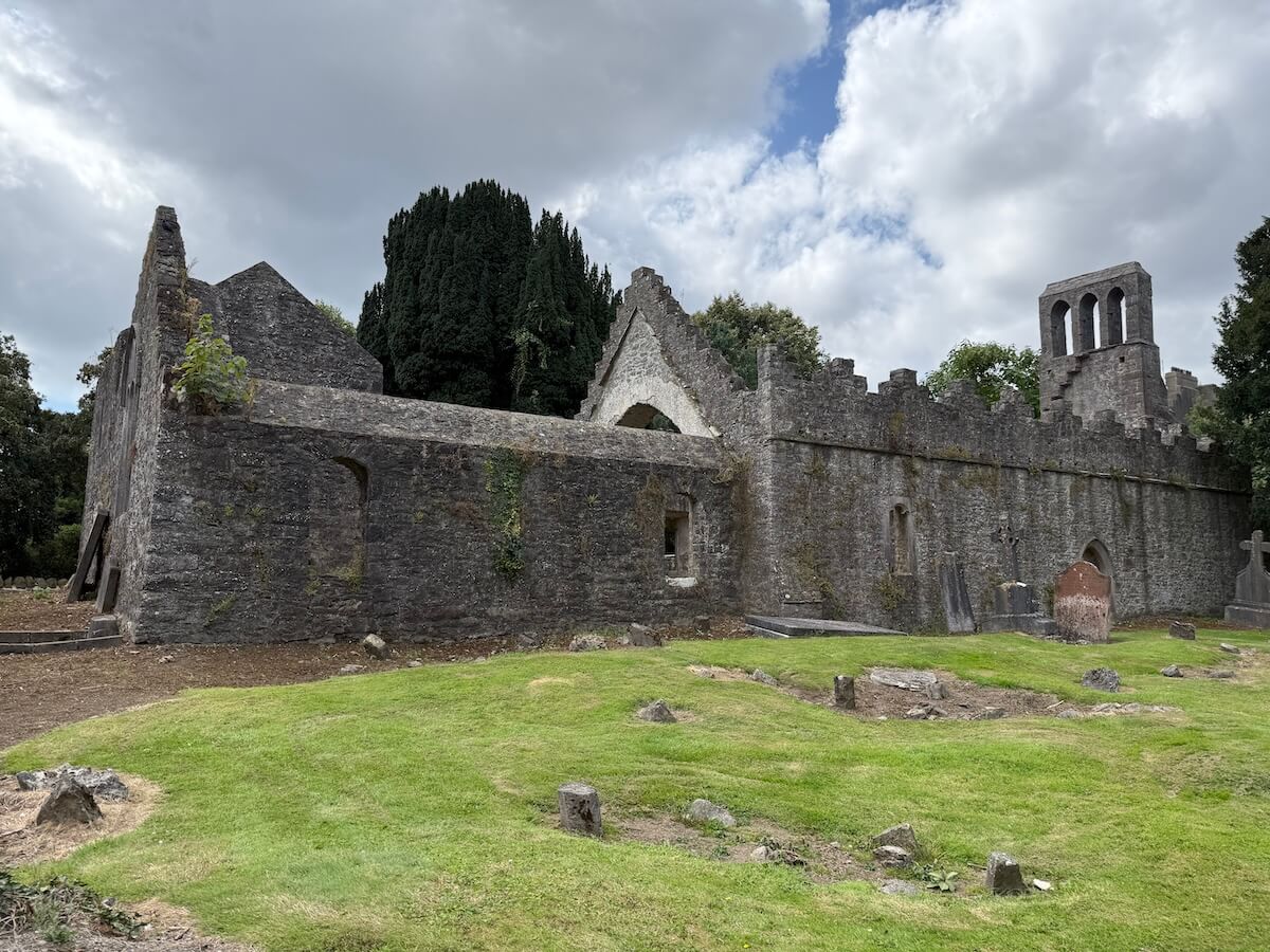 Stone ruins of an old medieval church stand under a cloudy sky, with arched windows and a bell tower partially intact. The grassy foreground shows remnants of graves and uneven stones, giving the site a quiet, historic feel.