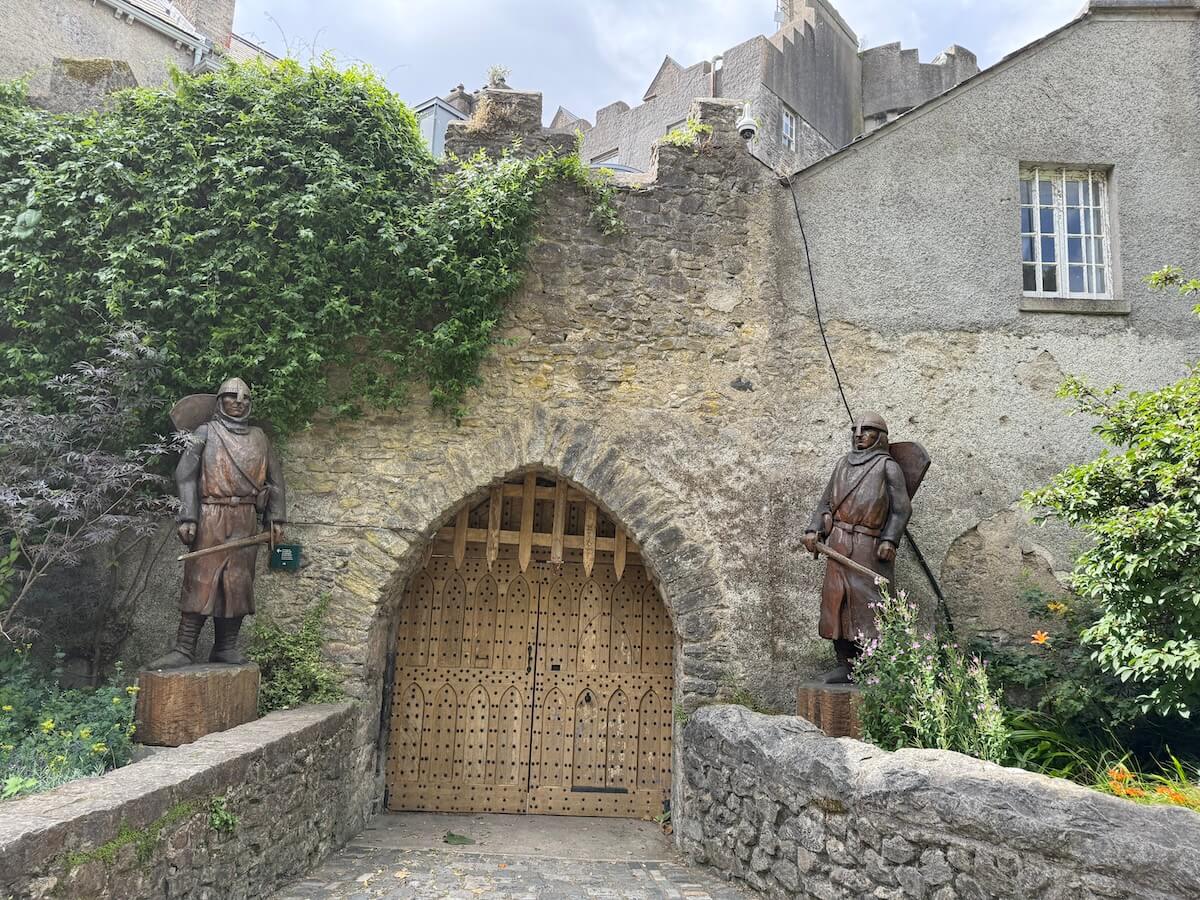 A medieval-style wooden gate set in a stone archway is flanked by two life-sized statues of armoured guards holding swords and shields. The weathered stone wall is partially covered in ivy, with a grey building and castle-like structure visible above.