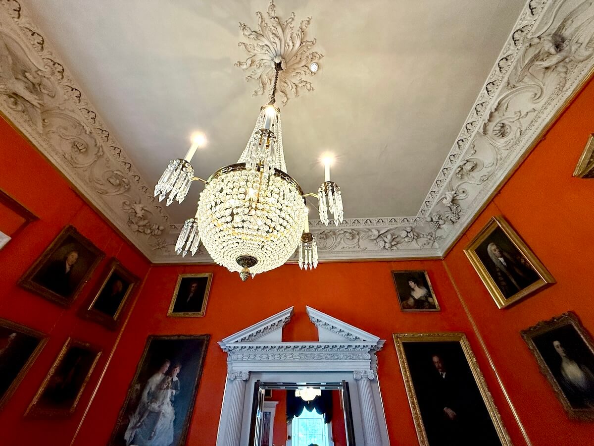 Crystal chandelier and ornate plaster ceiling in a richly decorated room at Malahide Castle, surrounded by framed portraits on orange walls.