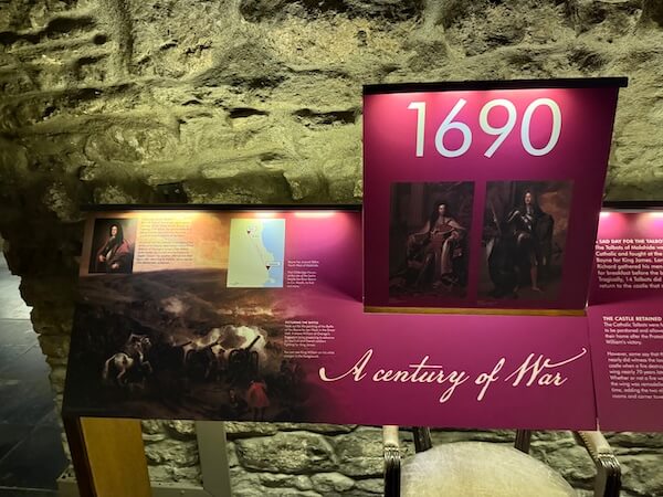 Museum panel titled “1690 – A Century of War” features portraits of two monarchs and a dramatic painting of a cavalry battle, set against a deep red background beneath a rough stone wall.