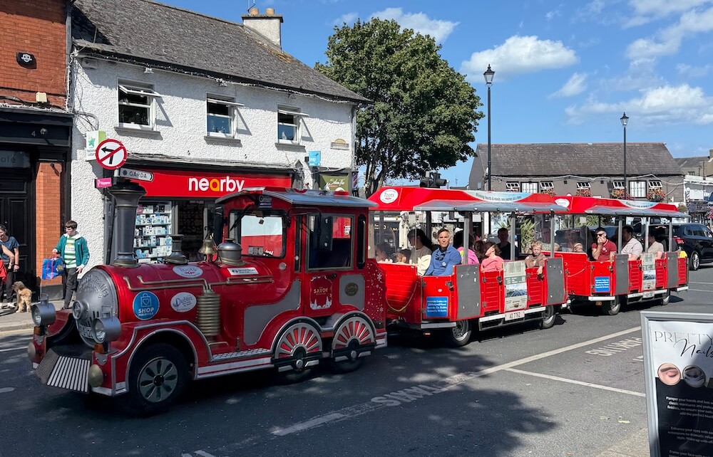 A bright red tourist train with open carriages full of passengers drives through a charming town center, passing shops and pedestrians on a sunny day. The train has a vintage-style engine and is marked with signs for local attractions and businesses.