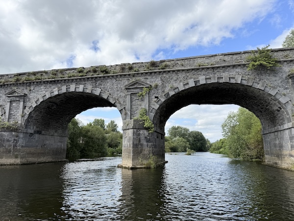 A stone arched bridge spanning a calm river, with greenery growing along its sides and cloudy blue sky above.