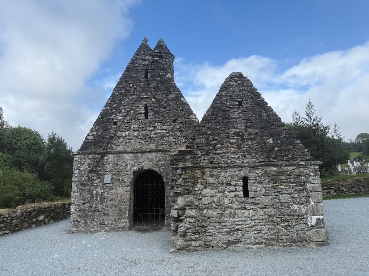 A stone medieval church with steeply pitched triangular gables stands against a partly cloudy sky. The weathered structure has narrow slit windows, a gated entrance, and is surrounded by gravel paths and low stone walls.