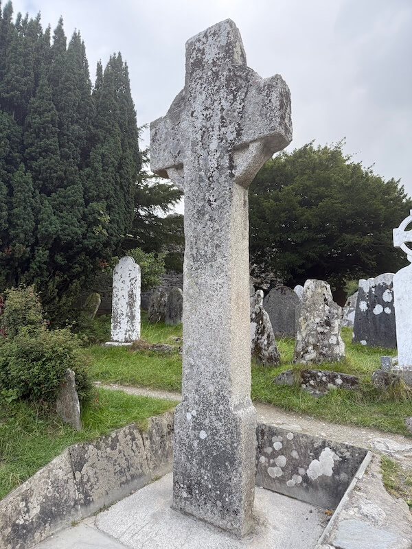 tall stone Celtic cross rises prominently in a graveyard, surrounded by smaller headstones and shaded by dense green trees.
