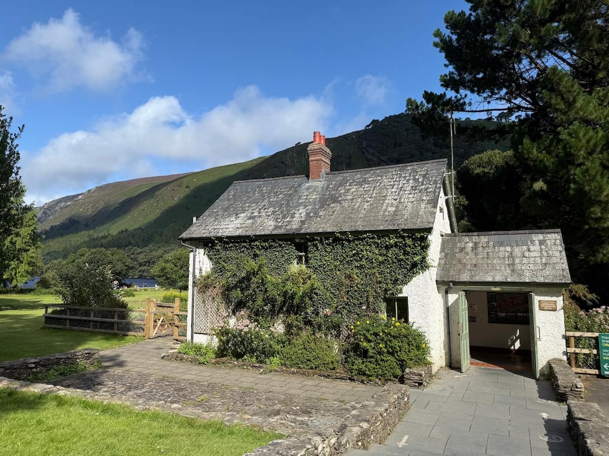 A quaint white stone cottage with a slate roof and ivy climbing its walls sits in a mountain valley, with a lake and rolling green hills in the background.