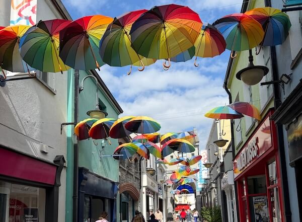A narrow street decorated with rows of colourful umbrellas hanging overhead, casting shade on the shops and pedestrians below.