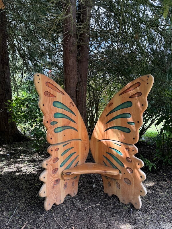A wooden bench shaped like butterfly wings, painted with green and orange accents, sits shaded beneath tall trees in a forested area.