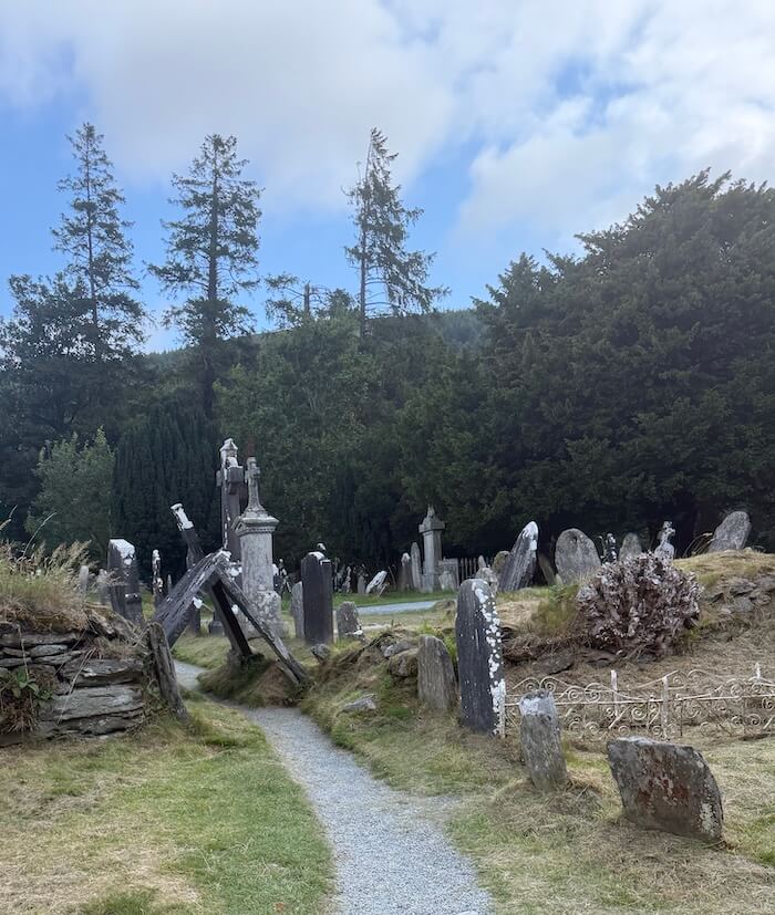 A narrow gravel path winds through an old graveyard with leaning headstones, surrounded by tall pine trees under a partly cloudy sky.