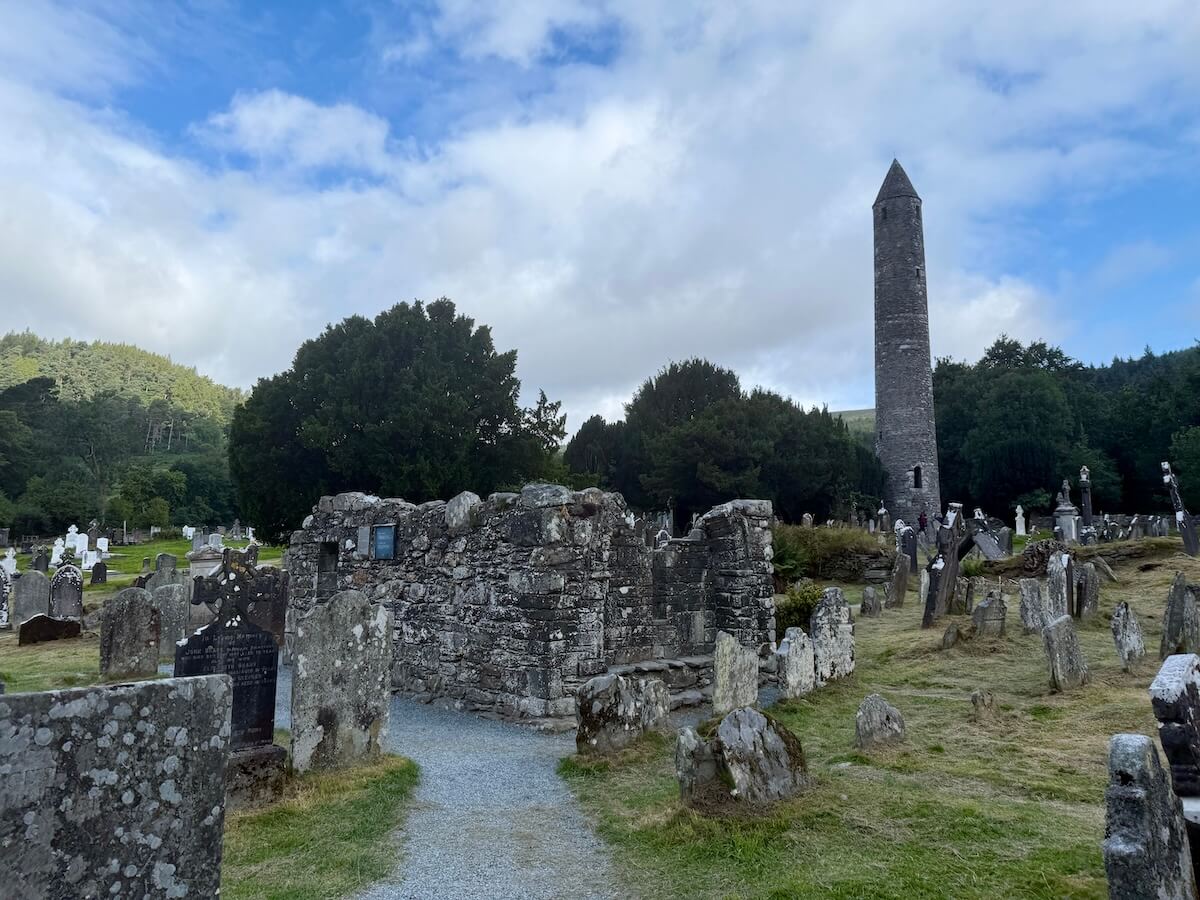 A historic stone graveyard features weathered headstones and the ruins of an old church, with a tall round tower rising in the background. Lush green trees and hills surround the site under a partly cloudy blue sky.