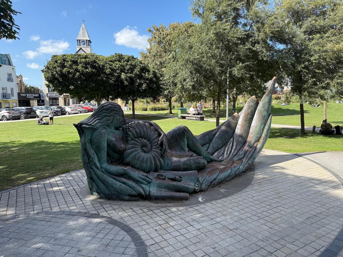 A bronze sculpture of a reclining mermaid cradling a large nautilus shell rests on a circular tiled platform in a grassy public park, with people relaxing on benches and a church clock tower visible in the background.