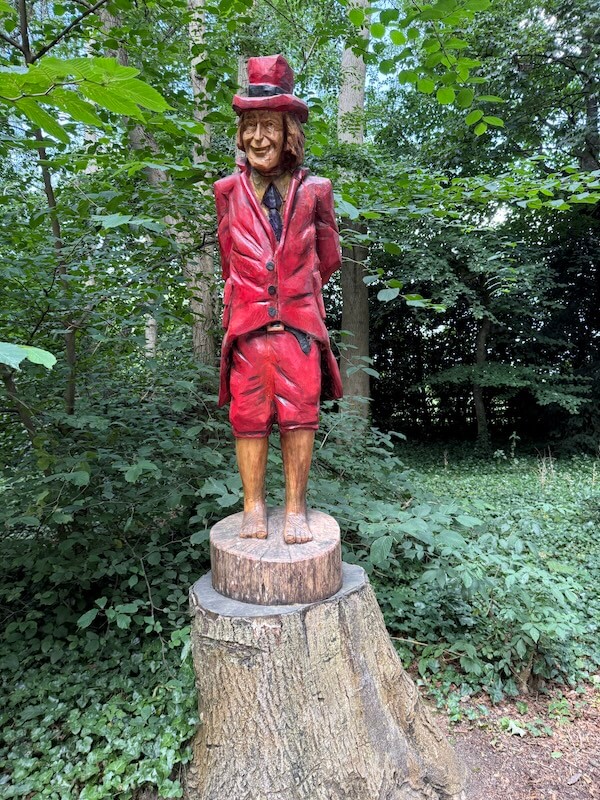 A whimsical wood-carved statue of a smiling man in a red suit and top hat stands on a tree stump among dense forest greenery.