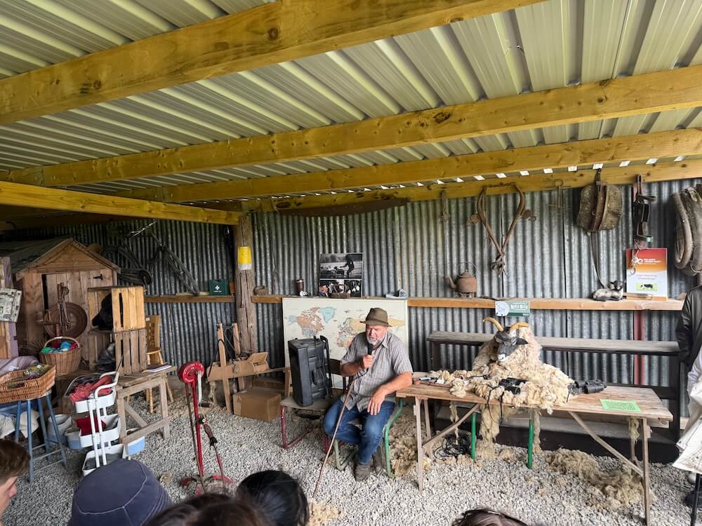 A farmer sits under a tin-roofed shed giving a demonstration with sheep’s wool on a table beside him, surrounded by old tools, farm equipment, and a world map pinned to the wall.
