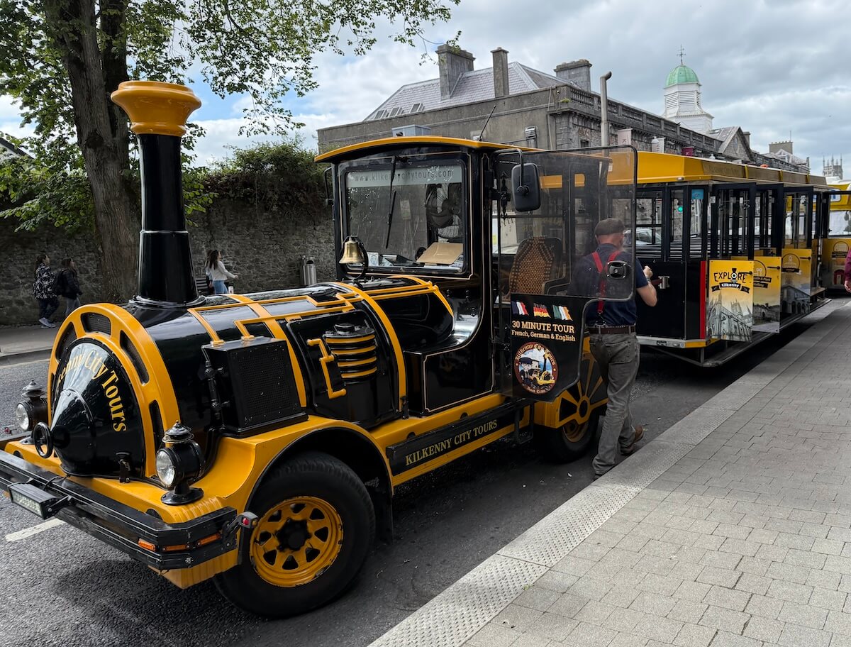 A black and yellow road train for Kilkenny City Tours is parked on a street, with a driver preparing the carriages for passengers.