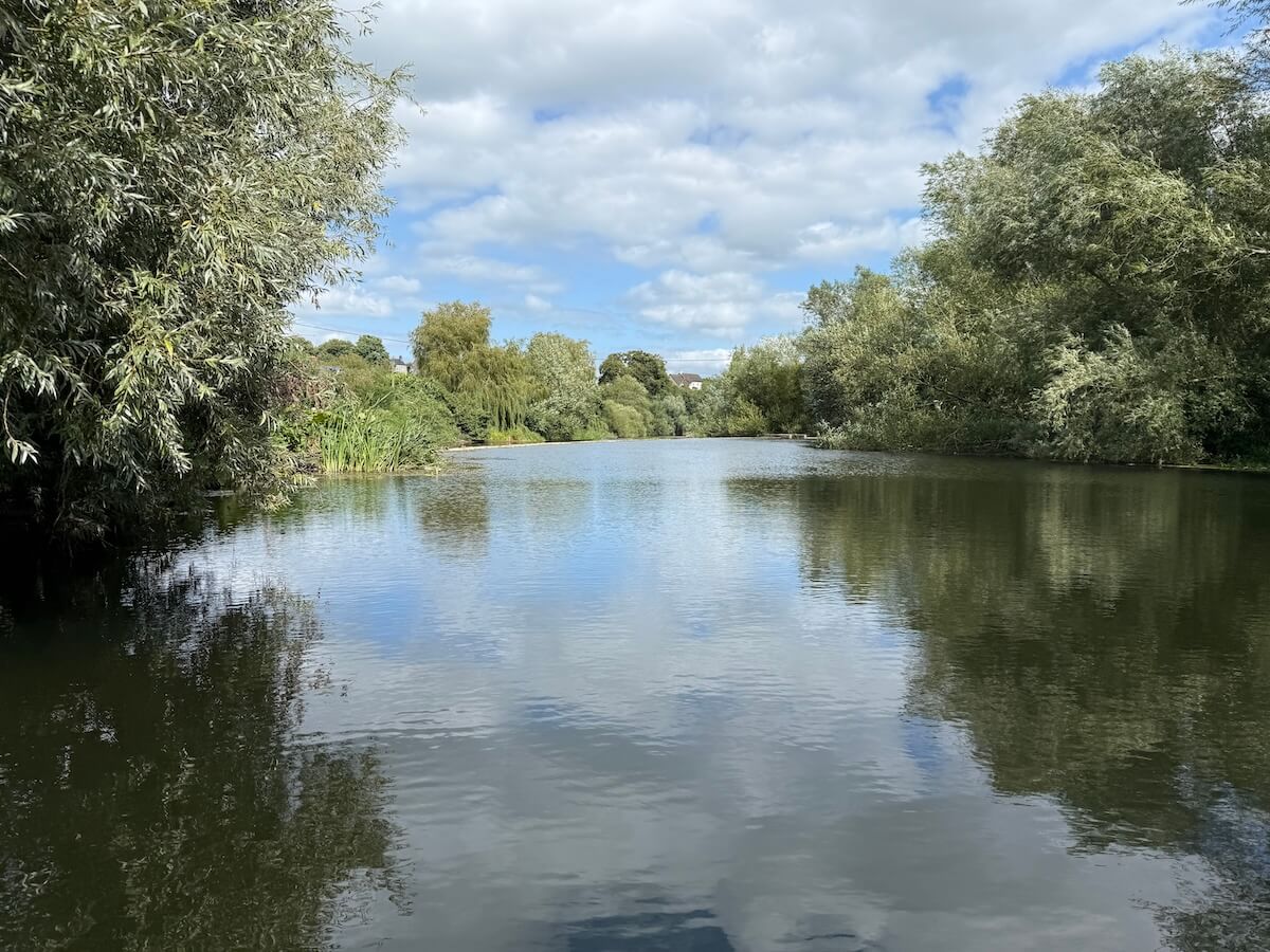 A wide, peaceful river bordered by leafy trees, their reflections mirrored on the still water under a partly cloudy sky.