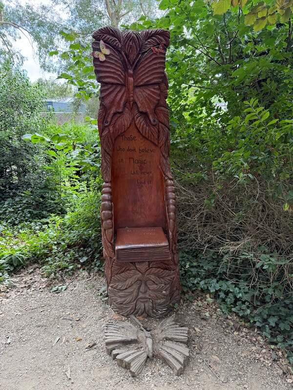 A tall, ornately carved wooden chair features butterfly and flower motifs, with engraved text that reads, “Those who don’t believe in magic will never find it,” surrounded by leafy forest plants.