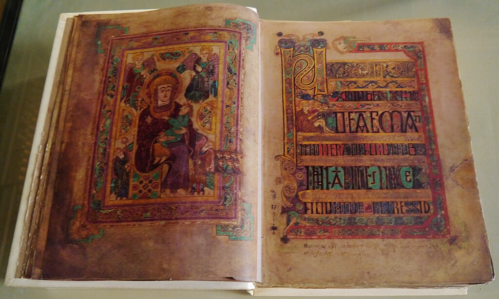 Open pages of the Book of Kells on display, featuring a richly coloured illustration of the Virgin and Child on the left and a highly ornate text page on the right with intricate Celtic knotwork, stylised letters, and vivid use of gold, green, and purple pigments.