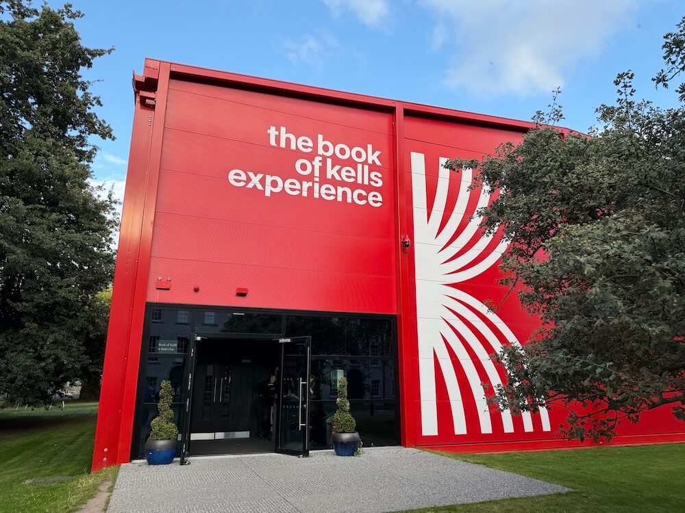 Bright red exterior of the Book of Kells Experience building featuring bold white lettering and a stylised design inspired by illuminated manuscripts, with glass entrance doors and manicured greenery on either side.