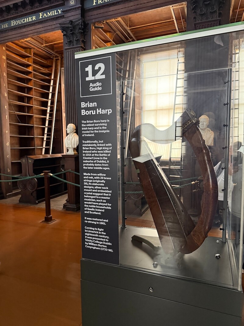 Display case containing the historic Brian Boru Harp at Trinity College Library, an ornately carved medieval instrument made of oak and willow, showcased with an audio guide panel explaining its origins and significance.