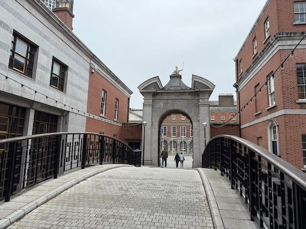 Curved stone footbridge leading to a historic arched gateway flanked by red-brick and grey-stone buildings, with two people walking beneath the arch on an overcast day.
