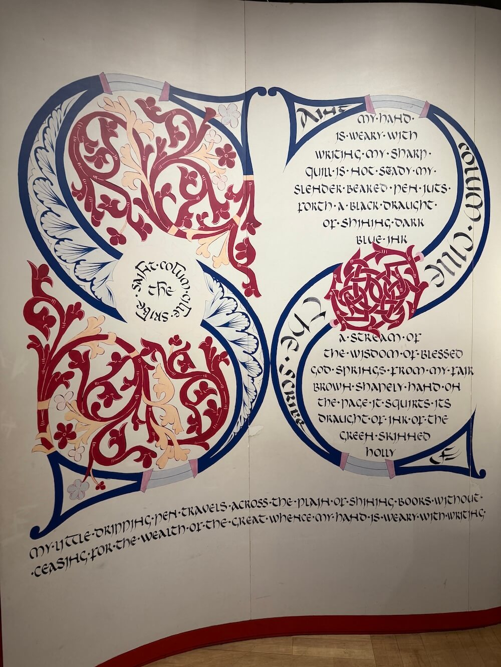 Wall display featuring a modern artistic interpretation of insular script with elaborate red and blue decorative initials and poetic text about the act of writing, styled to echo medieval manuscript tradition.