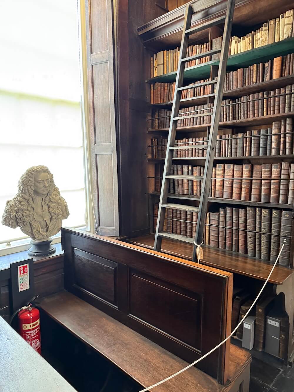 Interior corner of Marsh's Library featuring a wooden bench, a tall rolling ladder leaning on high bookshelves, and a marble bust next to a bright window.