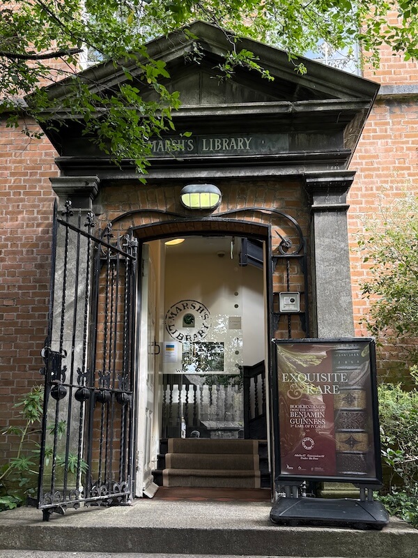 Exterior entrance of Marsh's Library in Dublin, showing a black iron gate, red brick facade, and a poster advertising a book exhibition titled “Exquisite & Rare.”