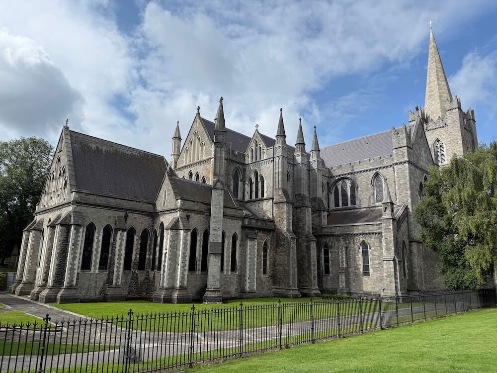 ide angle of St Patrick's Cathedral highlighting its pointed arches, stone buttresses, and intricate detailing, set against a partly cloudy blue sky and enclosed by a black wrought-iron fence.