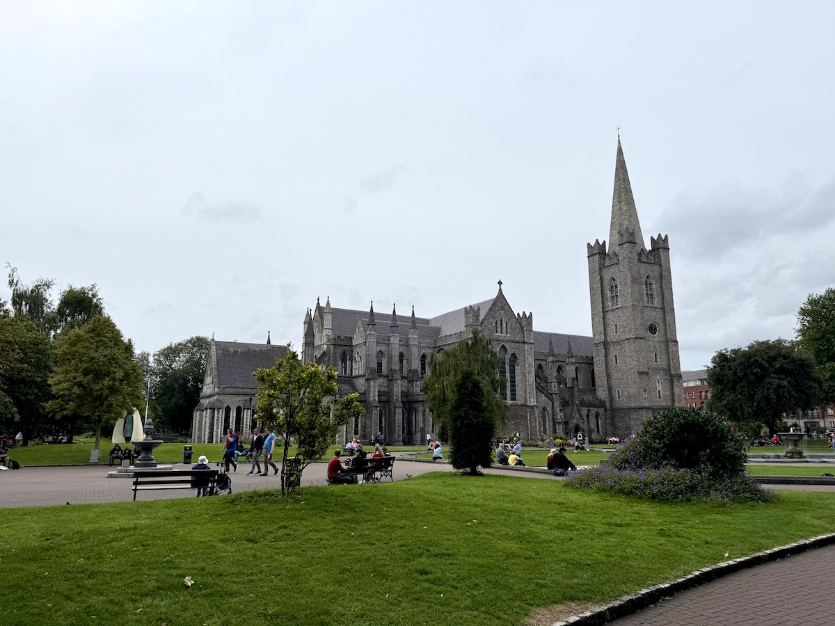 Wide view of St Patrick's Cathedral in Dublin, Ireland, showing its impressive Gothic architecture and tall spire under an overcast sky, with people relaxing in the surrounding landscaped park.