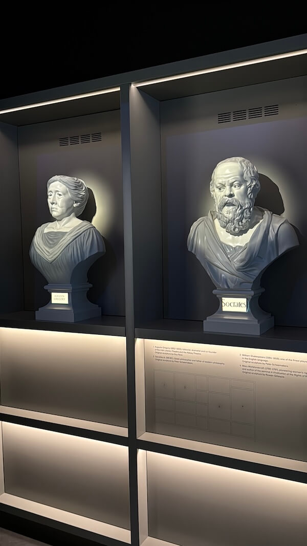 Marble busts of Augusta Gregory and Socrates displayed side by side in a dark grey modern exhibit case, each labelled with soft lighting enhancing the sculptural details.